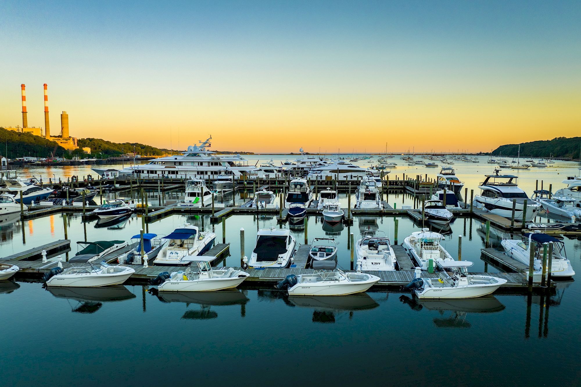 A marina with numerous boats docked, calm waters, a clear sky, and a coastal backdrop with tall smokestacks is in the distance at sunset.