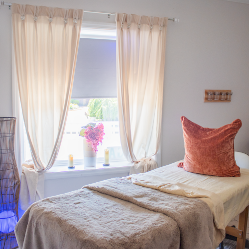 A cozy room with a massage table, soft pillows, and relaxing decor, including a vase and candles near a window with sheer curtains.