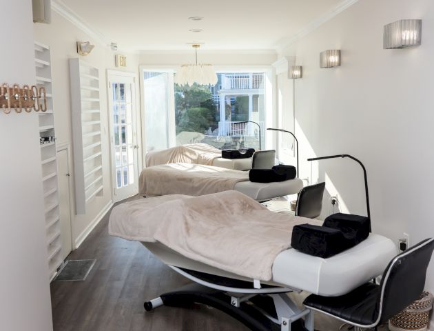 A modern, well-lit room with two spa or massage beds, blankets, chairs, and equipment, all arranged neatly on wood flooring.