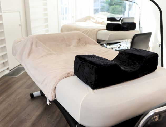 The image shows a clean, modern beauty or medical treatment room with a comfortable treatment bed, overhead lamp, and a padded head support.