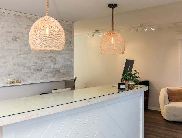 A modern reception area with a white counter, pendant lights, and a cozy seating area. A computer and plant are on the counter.