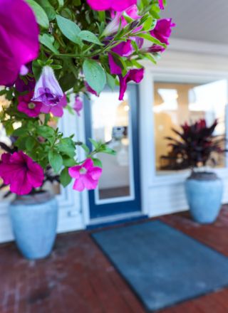 This image shows a porch with flowering plants in pots, a blue door, and a welcome mat. Bright pink flowers are in the foreground.