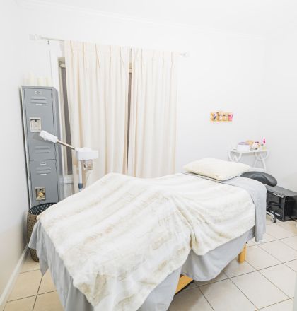 A small, well-lit massage or treatment room with a bed, blanket, medical equipment, and calming decor is shown.