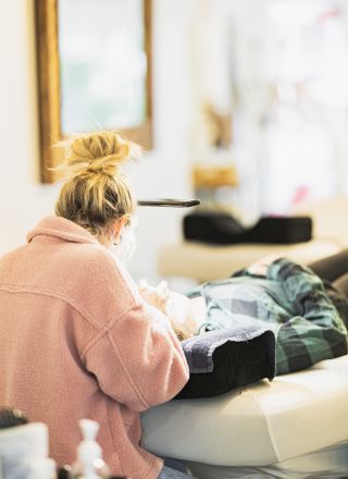 A person wearing a pink sweater and a top knot hairstyle works on eyebrow grooming for a client reclining on a salon chair in a bright room.