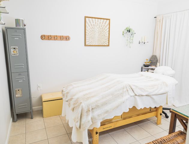 A cozy room with a massage table, soft blanket, gray locker, wooden bench, wall art, small table with a lamp, and light-colored curtains.