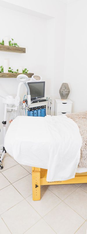 The image shows a clean, well-lit treatment room with a bed covered in soft, beige blankets, shelves with plants, and various spa equipment.