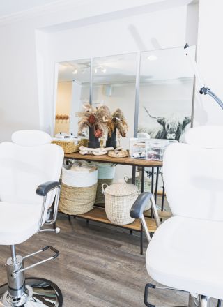 The image shows a bright room with two white salon chairs, a wall mirror, various decor items, and beauty products on a shelf.