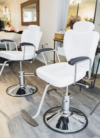 The image displays a modern salon interior with two white barber chairs, mirrors, and a person in the background.