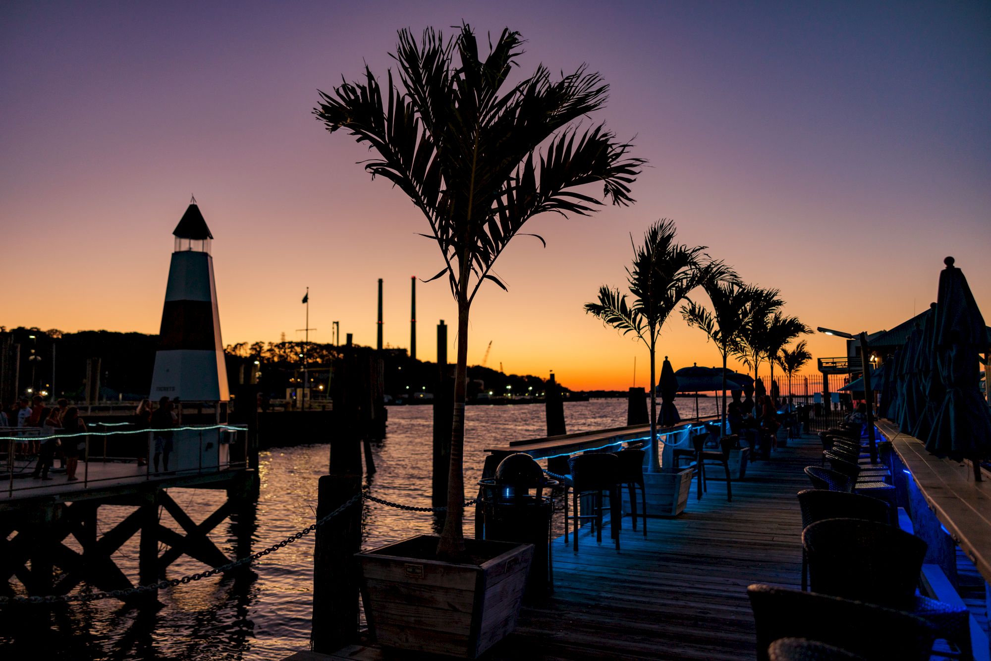 Sunset view of a waterfront with palm trees, a lighthouse, and outdoor seating on a wooden deck.