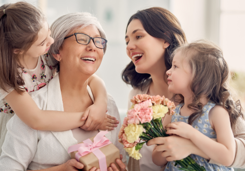 A joyful family scene with a grandmother holding a gift, surrounded by two young girls and a woman, all smiling and sharing a happy moment together.