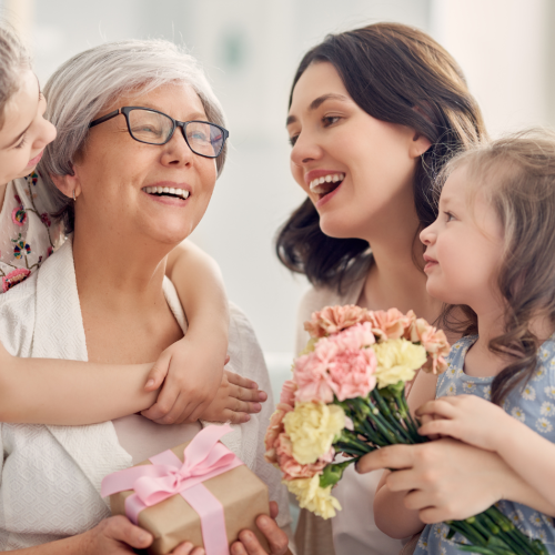 A joyful family scene with a grandmother holding a gift, surrounded by two young girls and a woman, all smiling and sharing a happy moment together.