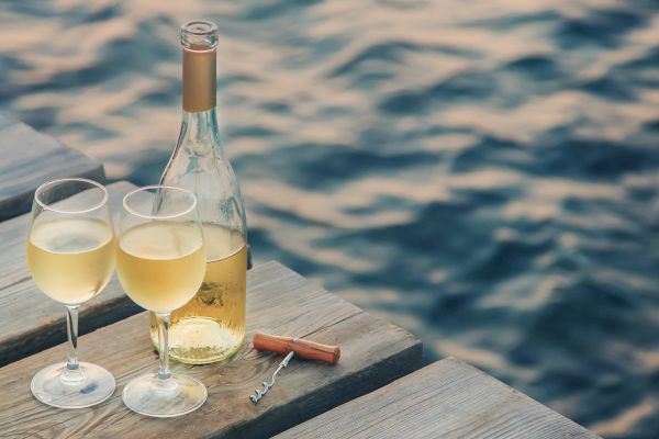 A bottle of white wine and two filled wine glasses sit on a wooden dock overlooking the water, accompanied by a corkscrew.