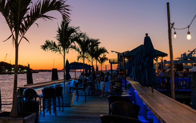 An outdoor bar or restaurant by the water at sunset, featuring palm trees, seating areas, string lights, and people enjoying the evening atmosphere.