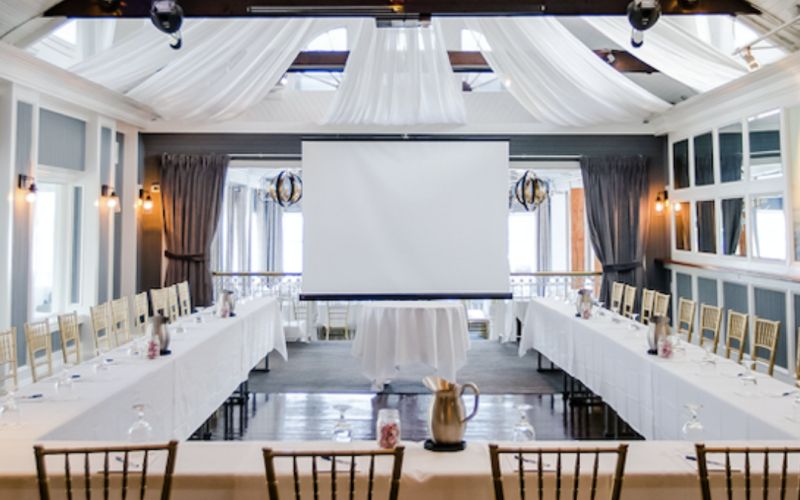 A well-lit conference room with a U-shaped table setup, chairs, a white screen, and elegant white drapes on the ceiling.