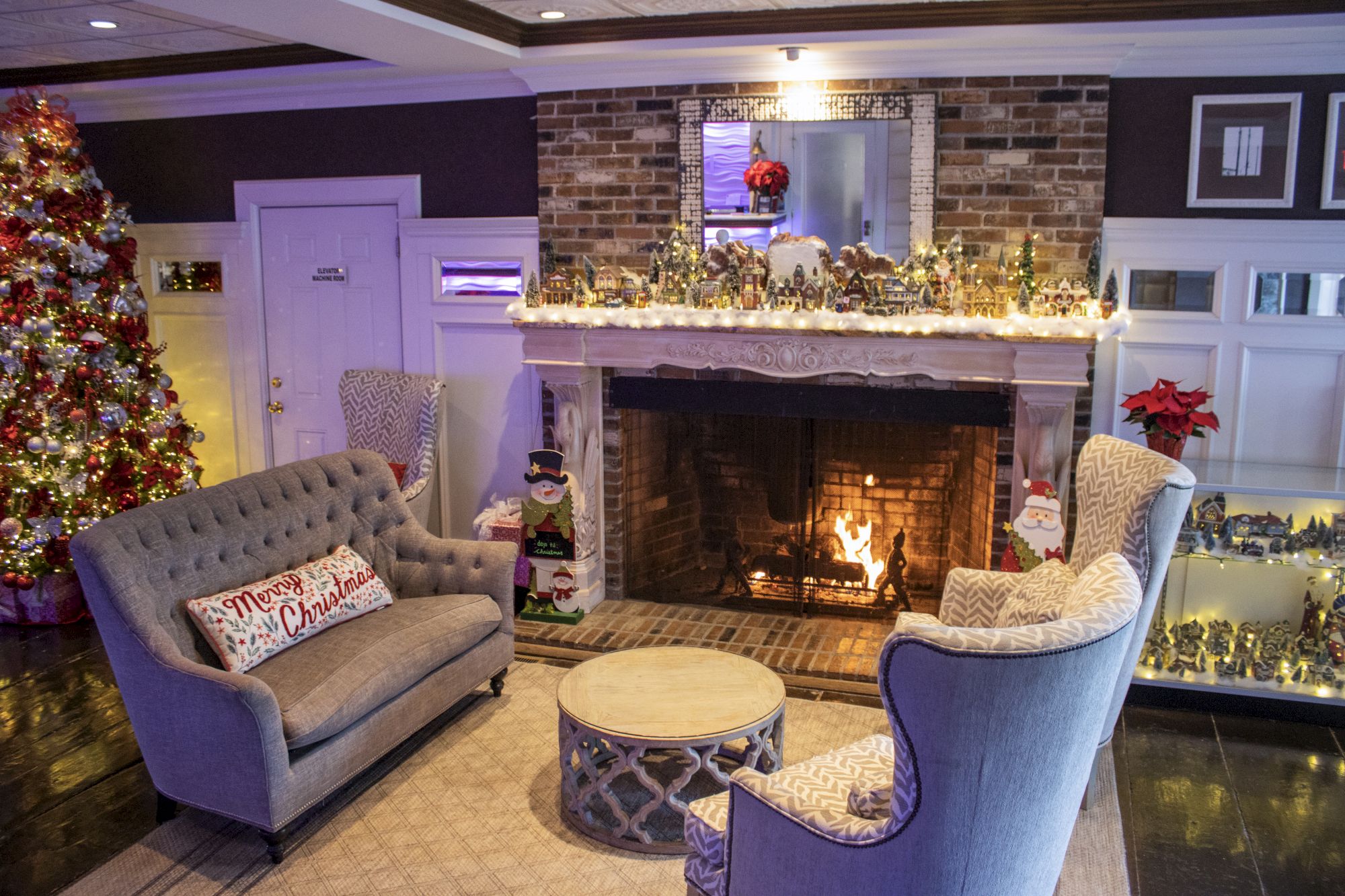 A cozy living room with a Christmas tree, lit fireplace, armchairs, festive decorations, and snowman figures sits warmly decorated for the holidays.