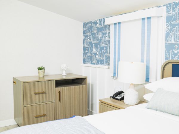 The image shows a bedroom with nautical-themed wallpaper, a bed, a nightstand, a lamp, a phone, and a dresser with decor items.
