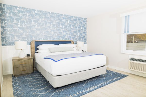 The image shows a bedroom with a bed, two nightstands, lamps, nautical-themed wallpaper, a blue rug, and a window with an air conditioner.