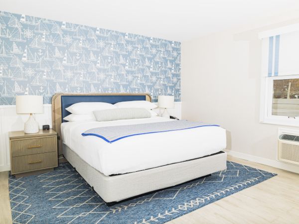 The image shows a bedroom with a bed, two nightstands, lamps, nautical-themed wallpaper, a blue rug, and a window with an air conditioner.