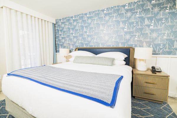 A bedroom with a nautical-themed wallpaper, a neatly made bed, and two bedside tables with lamps, telephone, and a light blue blanket.