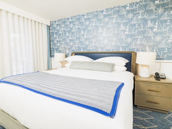 A bedroom with a nautical-themed wallpaper, a neatly made bed, and two bedside tables with lamps, telephone, and a light blue blanket.