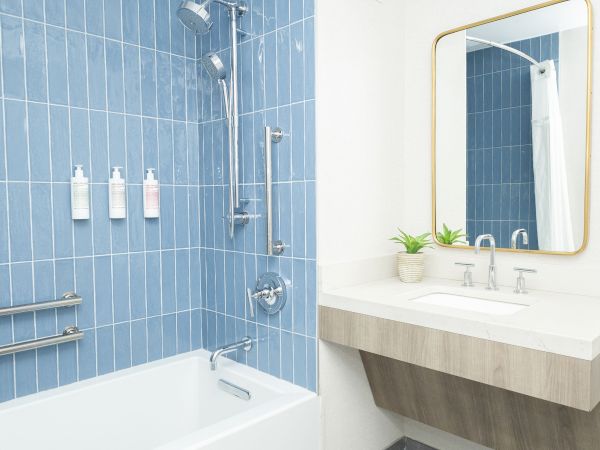 A modern bathroom with a blue tiled shower, rainfall head, soap bottles, and a white sink with faucets and mirror.