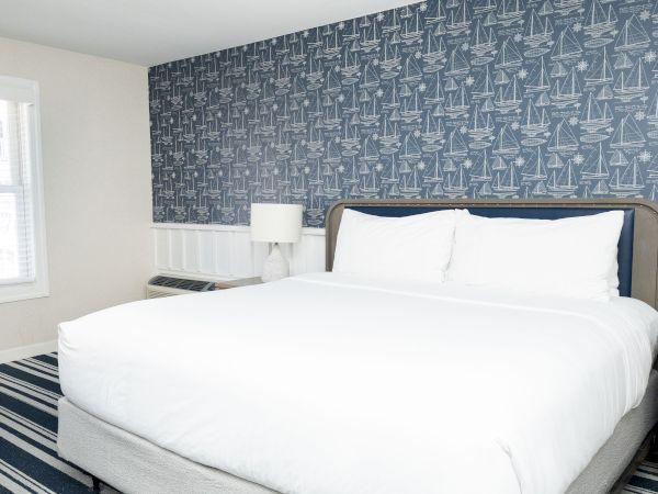 A bright bedroom with a white bed and navy wallpaper covered in sailboats and nautical charts, by a window.