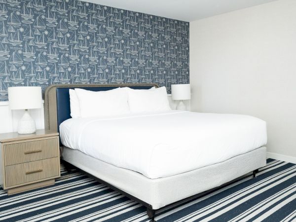 A bright bedroom with a large white bed, striped rug, and blue nautical sailboat wallpaper covering the wall.