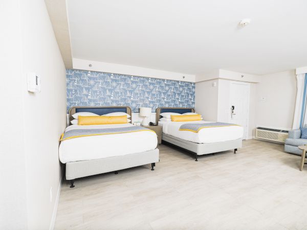 A hotel room with two beds, blue wallpaper, light wood flooring, a couch, and a small table. Bright, modern, and minimalistic design.