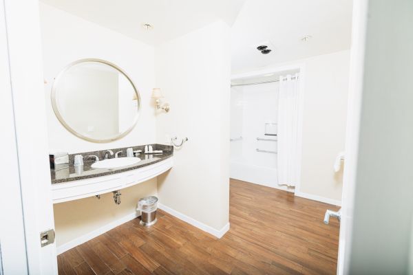 The image shows a clean bathroom with a round mirror, sink, soap dispenser, trash can, wood flooring, and shower area with a curtain.