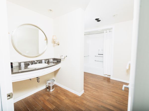 The image shows a clean bathroom with a round mirror, sink, soap dispenser, trash can, wood flooring, and shower area with a curtain.