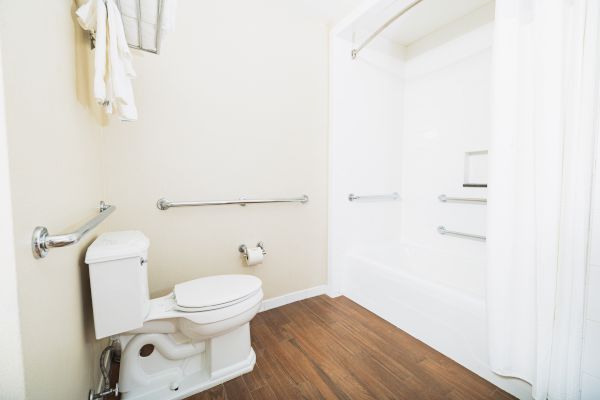 A bathroom with a toilet, bathtub, shower curtain, grab bars, wooden floor, and towels hangs beside the toilet.