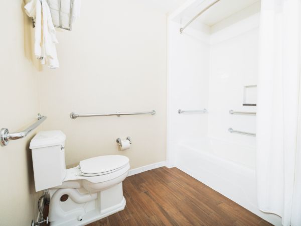 A bathroom with a toilet, bathtub, shower curtain, grab bars, wooden floor, and towels hangs beside the toilet.