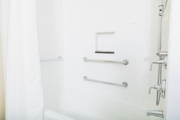 The image shows a white bathtub with a shower, three grab bars, a built-in shelf, and a shower curtain in a tiled bathroom.