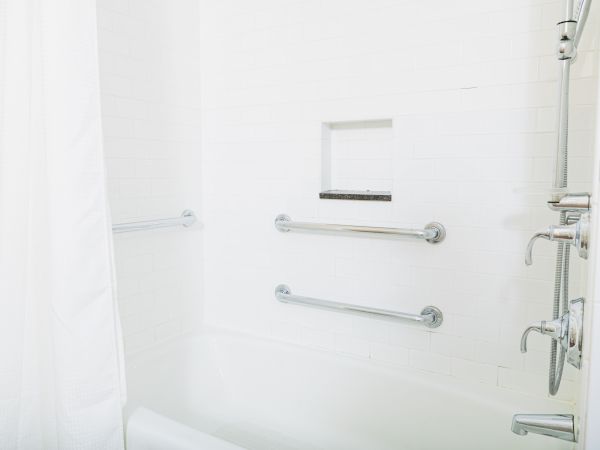 The image shows a white bathtub with a shower, three grab bars, a built-in shelf, and a shower curtain in a tiled bathroom.