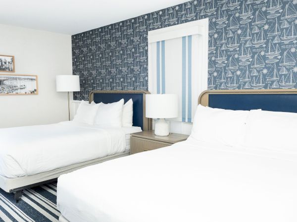 A bright nautical-themed bedroom with two beds, white linens, bedside lamps, and sailboat wallpaper covering the walls.
