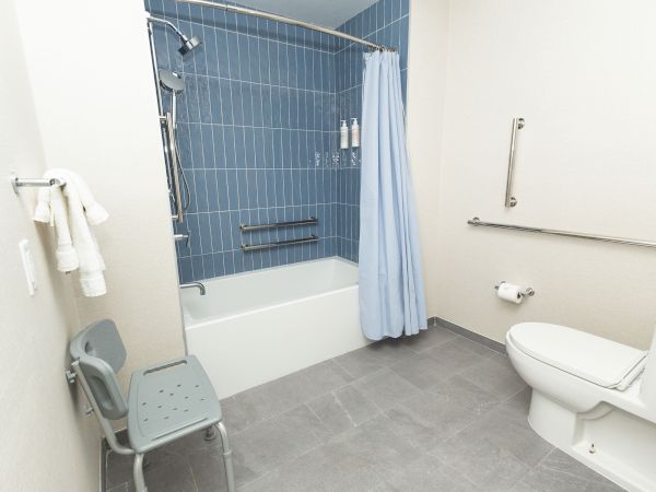 A clean accessible bathroom with a bathtub and shower curtain, towels, grab bars, a shower head, and an adjacent toilet.