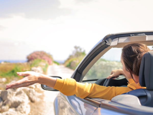 A person in a yellow shirt drives a convertible on a scenic road, with an arm outstretched, enjoying the view.