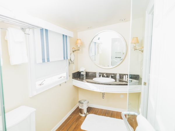A bright, modern bathroom with a round mirror, a washbasin with a granite top, white towels, and a small trash can on a wooden floor ending the sentence.