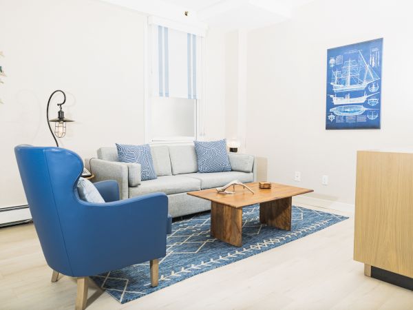A living room with a blue armchair, grey sofa, wooden coffee table, and nautical-themed decor on a blue rug near a window and a wall painting.