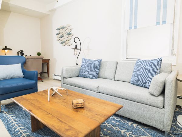 A modern living room with a gray sofa, blue armchair, wooden coffee table, and wall art. Blue and white accents adorn the space, with good lighting.