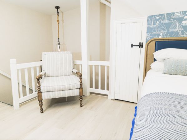The image shows a cozy, white-themed bedroom with nautical wallpaper, a striped armchair, and a bed with blue and white bedding.