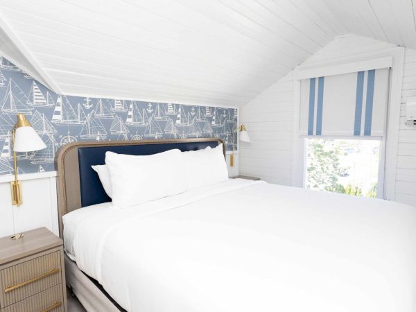 A bright nautical-themed bedroom with a large bed, white pillows, gold lamps, and sailboat wallpaper.