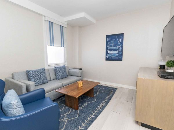 A modern living room with a gray sofa, blue armchair, wooden coffee table, patterned rug, window blinds, wall art, and a TV.