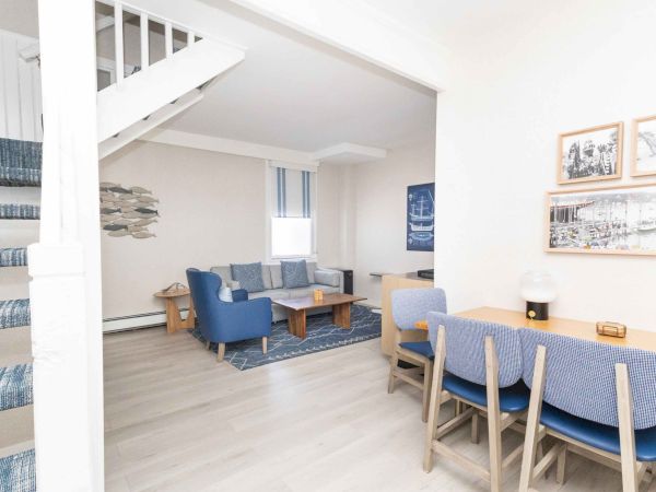 Bright nautical living room with blue sofas, wood tables, framed photos, and fish wall art under a white stairwell.