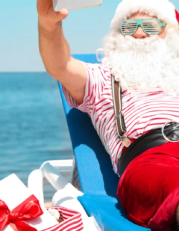 Santa Claus is lounging on a beach chair, taking a selfie, with a gift bag beside him under a sunny, clear sky.