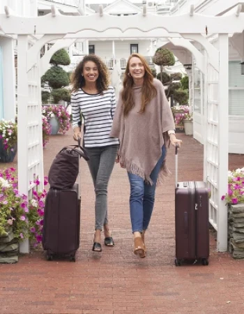 Two people walking with suitcases on a brick path surrounded by flowers and white buildings, smiling under a wooden archway.