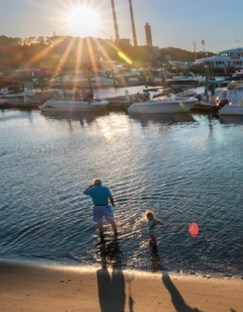 A person and a child stand in shallow water at sunset, with boats and an industrial backdrop in the distance.