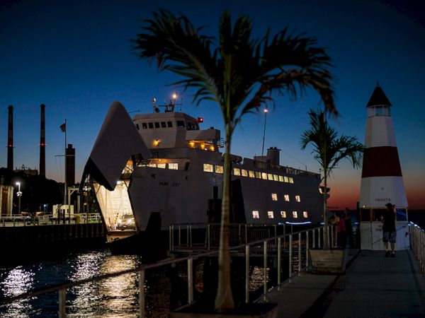 A docked ship is lit up at night, with palm trees and a lighthouse nearby, set against a deep blue evening sky.