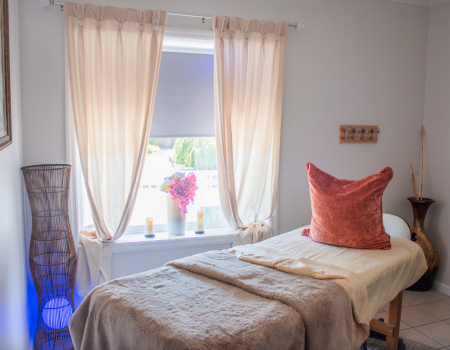 A cozy room with a massage table, soft lighting, curtains, and decorative pillows, creating a relaxing atmosphere.