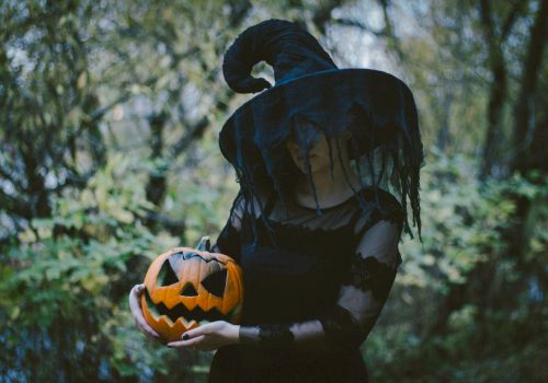 A person in a witch costume holds a carved pumpkin outdoors, surrounded by trees and foliage.